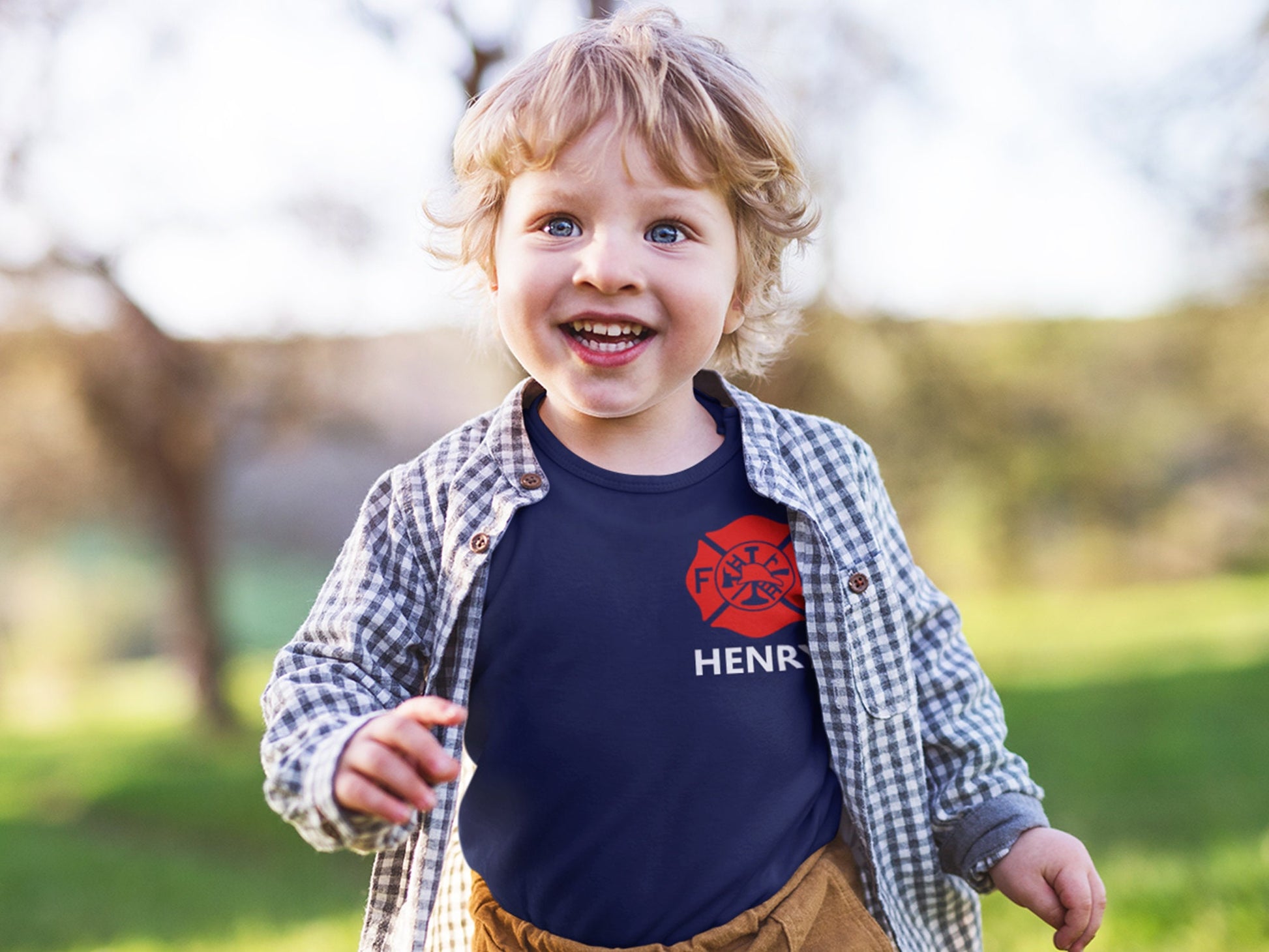 Toddler wearing the personalized firefighter baby bodysuit under a plaid shirt, showing the red Maltese Cross and custom name