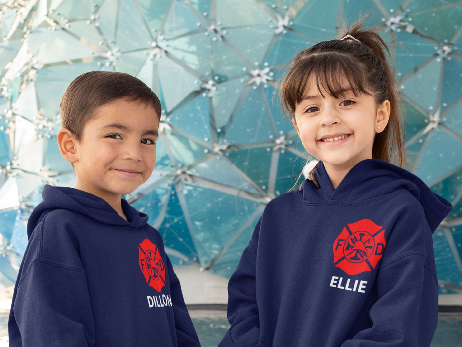 Two children wearing navy blue hoodies with a red firefighter logo and white text. The text on the hoodie on the left says 'Dillon' and the text on the hoodie on the right says 'Ellie' showing customization option.