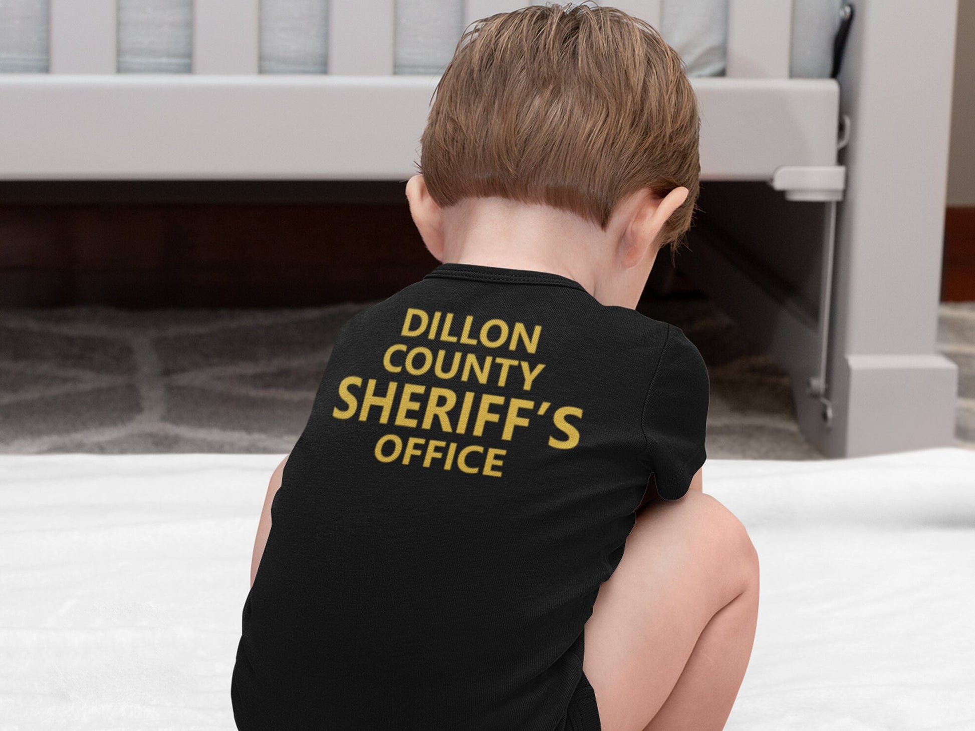 Squatting baby wearing a black short sleeve bodysuit. Back of bodysuit shows custom county sheriffs office in gold text.