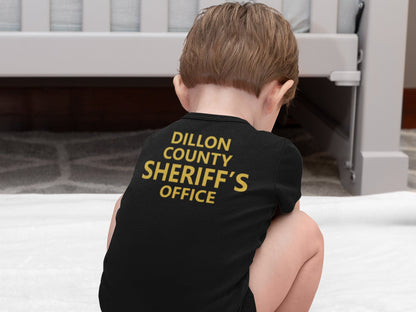 Squatting baby wearing a black short sleeve bodysuit. Back of bodysuit shows custom county sheriffs office in gold text.