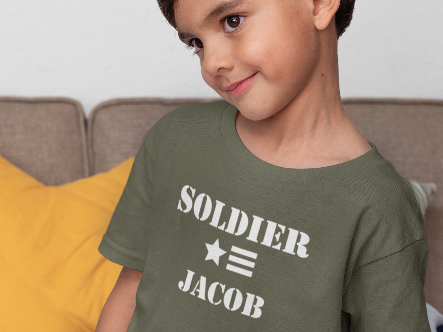 Boy wearing a green t-shirt with white text that says 'SOLDIER' with a star and three stripes underneath and 'JACOB' below the stripes.