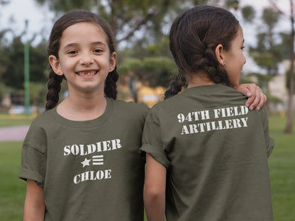 Twin girls wearing green t-shirts with white text. Front facing girl's shirt says 'SOLDIER' on the top line, a star and 3 stripes on the second line, and 'CHLOE' on the third line. Back facing girl's shirt says '94TH FIELD ARTILLERY' on the back.