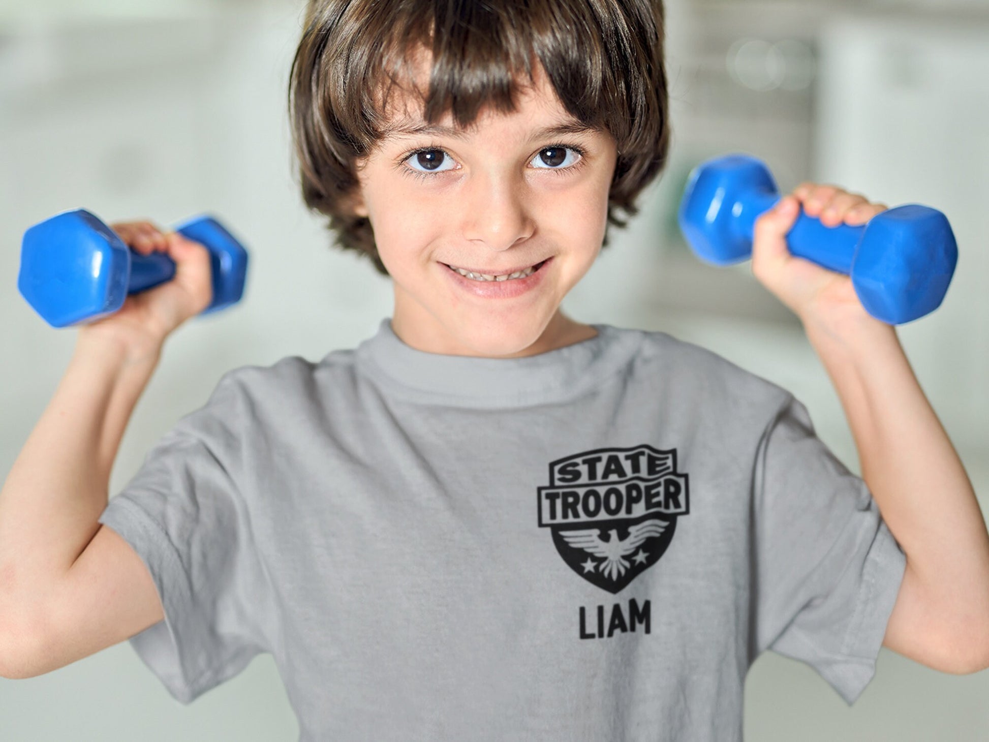 Boy wearing a gray short sleeve t-shirt with a black graphic of a shield with an eagle and the text 'STATE TROOPER' and 'LIAM' underneath in black text.
