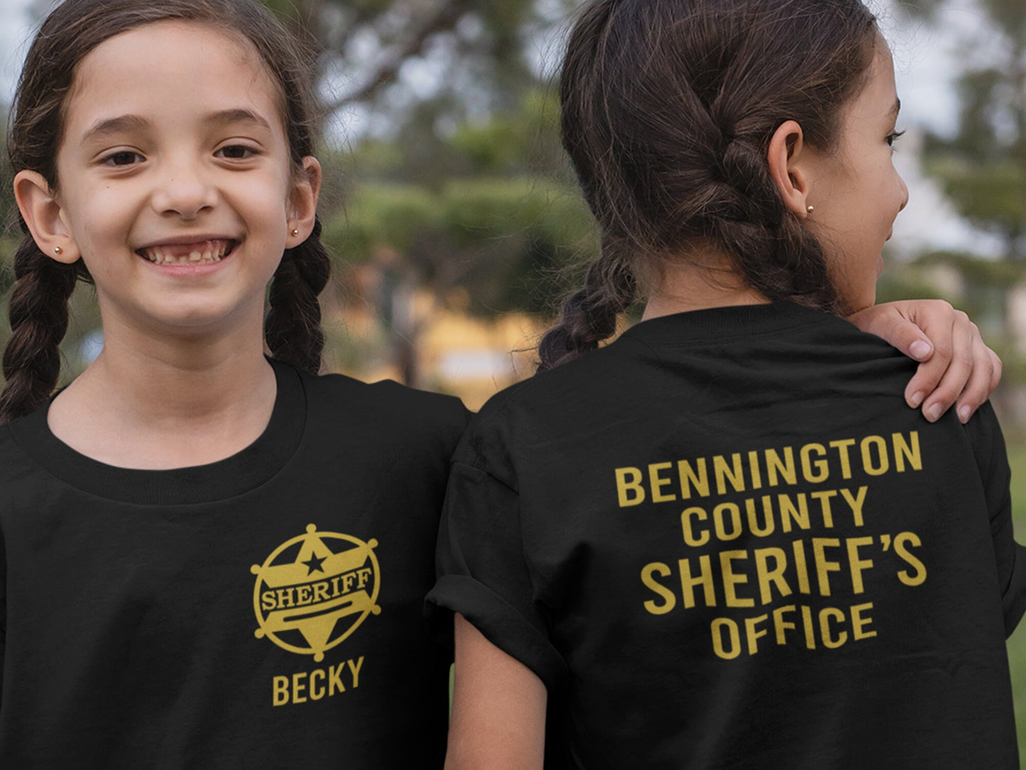 Twin girls, both wearing black t-shirts, one facing forward with a gold sheriff's badge on the left chest, and the name Becky underneath. The other girs is facing backwards with "Bennington County Sheriff's Office" in gold lettering on the back of the shirt.