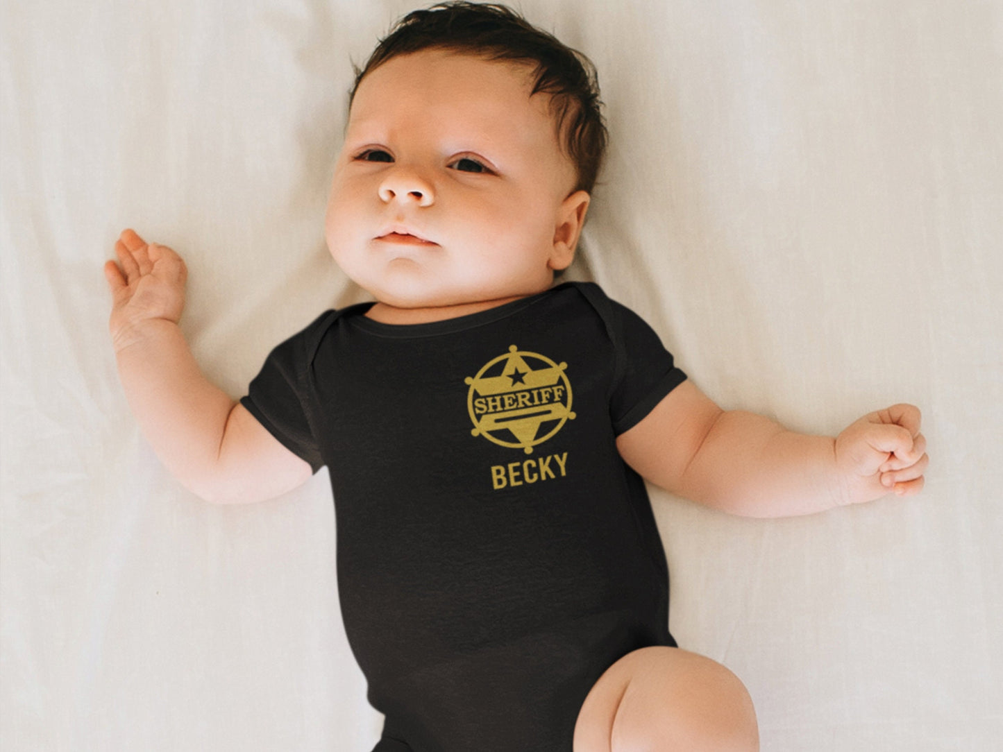 Baby laying in a crib wearing a black shower sleeve bodysuit with gold sheriff’s badge on left chest and personalized name underneath.