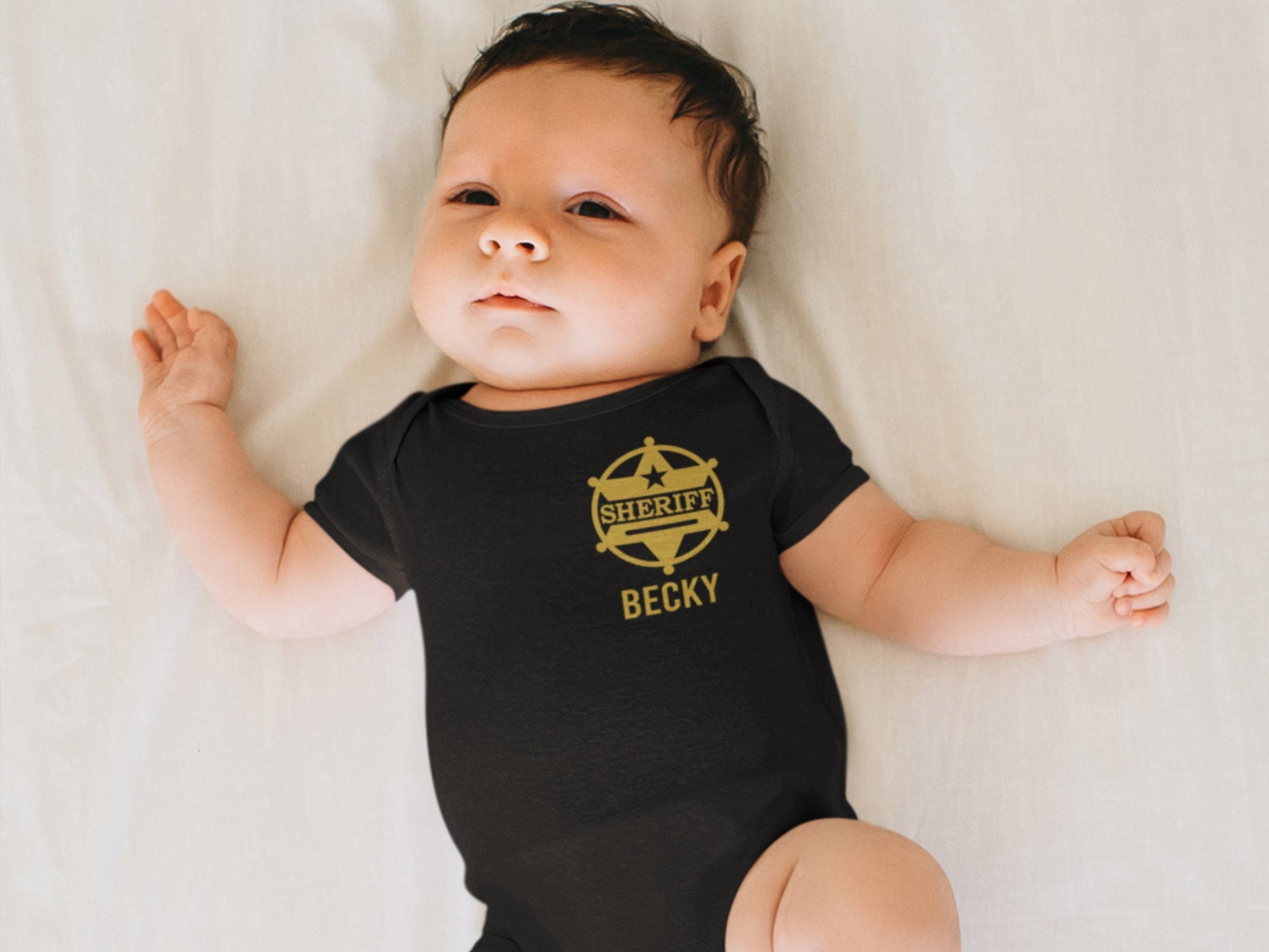Baby laying in a crib wearing a black shower sleeve bodysuit with gold sheriff’s badge on left chest and personalized name underneath.
