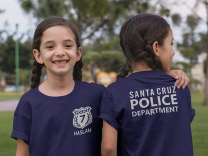 Twin girls wearing navy blue t-shirts. Girl on left showing the front with a silver police badge and number 7 and her name underneath. Left girl showing back of shirt with silver text "santa cruze police department".
