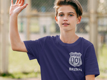 A purple t-shirt with a silver police badge design. The badge has the word 'POLICE' and the number '10' inside. The name 'BRENN' is printed below the badge.