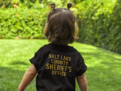 Toddler girl wearing a black tee facing backward. The shirt says Salt Lake County Sheriff's Office in gold letting.