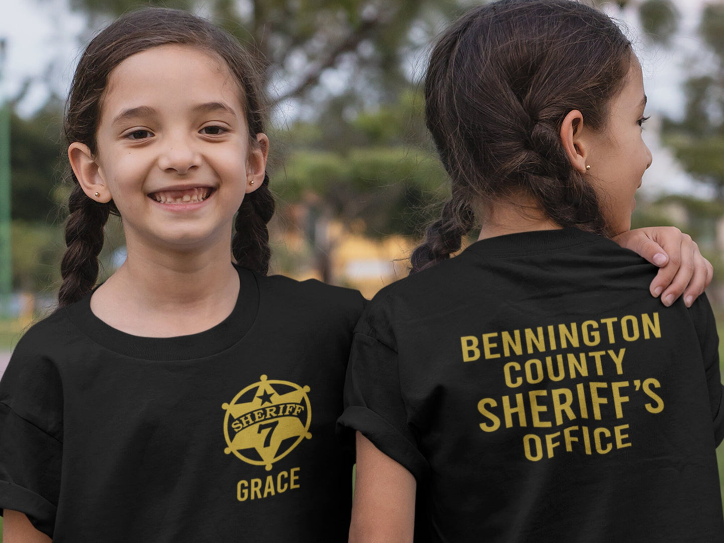 Twin girls wearing black t-shirt with a gold sheriff's star badge on the front and the text 'SHERIFF 7 GRACE' underneath. The back of the shirt has the text 'BENNINGTON COUNTY SHERIFF'S OFFICE' in gold lettering.