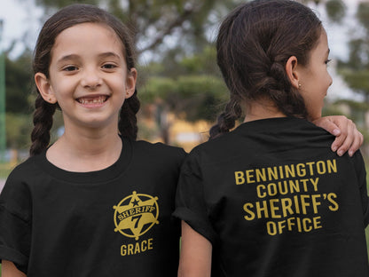 Twin girls wearing black t-shirt with a gold sheriff's star badge on the front and the text 'SHERIFF 7 GRACE' underneath. The back of the shirt has the text 'BENNINGTON COUNTY SHERIFF'S OFFICE' in gold lettering.