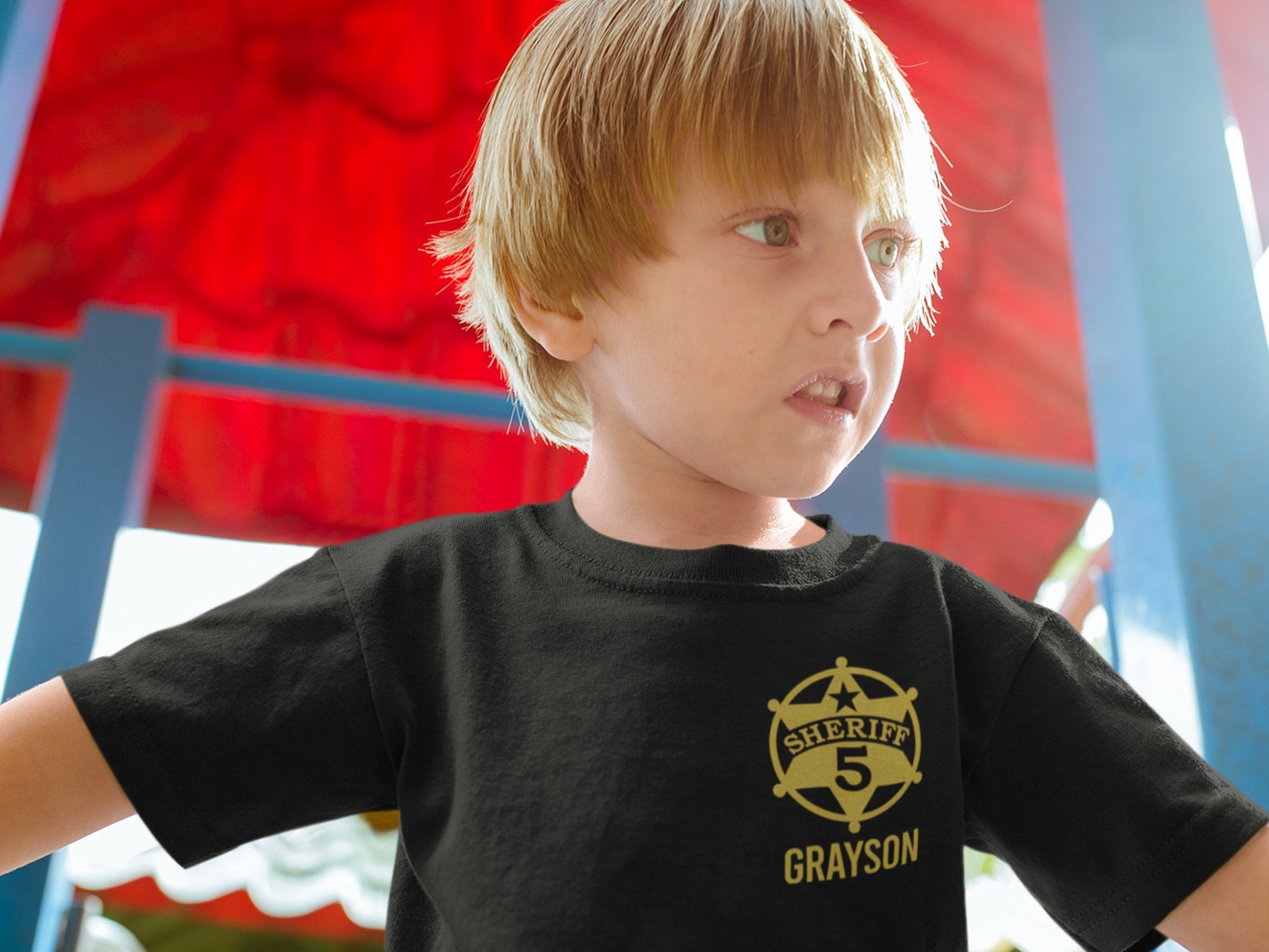 A child wearing a black t-shirt with a gold sheriff's badge that says 'Sheriff 5 Grayson'.