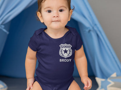 Toddler sitting wearing a navy blue short sleeve bodysuit with silver police badge and baby’s name underneath.