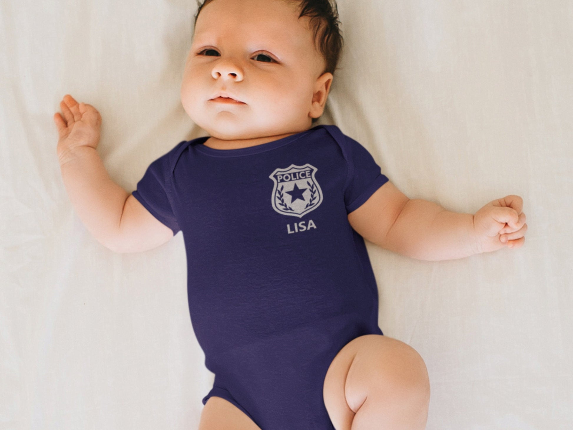Small baby in a crib wearing a short sleeve navy blue bodysuit with silver police badge and custom name underneath.