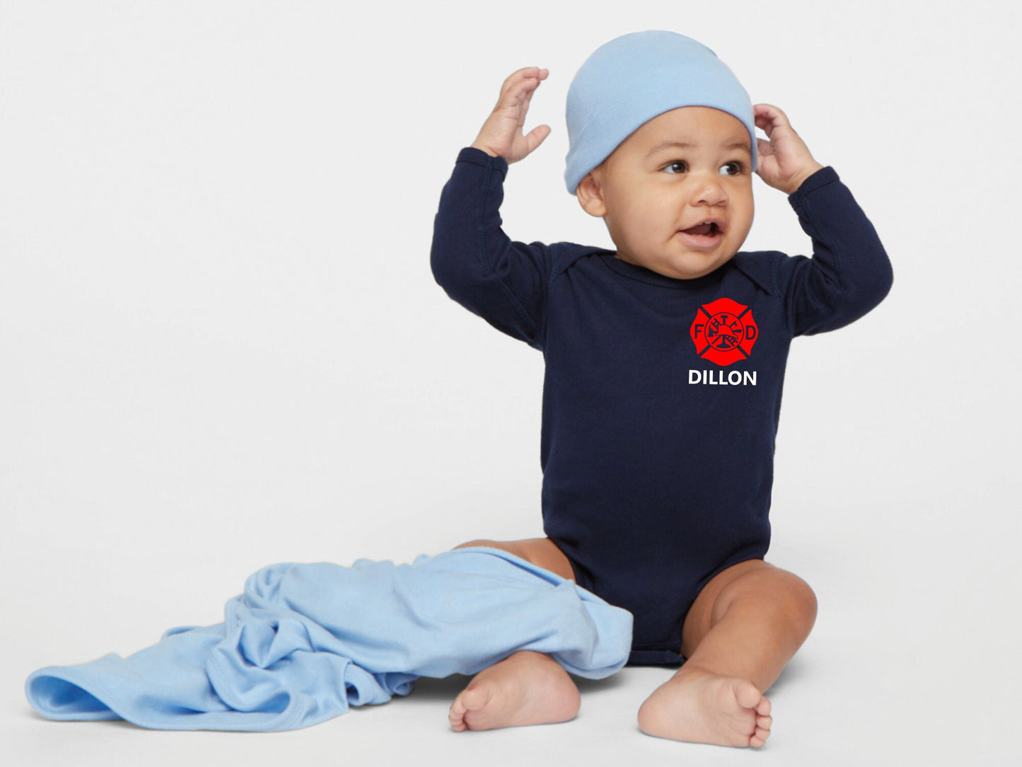 Sitting baby wearing a navy blue long sleeve bodysuit with red Maltese cross on less chest and name underneath. Also wearing a light blue knitted hat and light blue blanket on his leg.