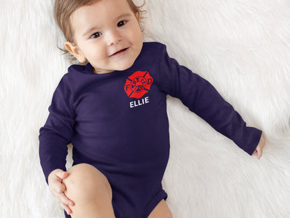 Baby in crib wearing a navy blue long sleeve bodysuit with red Maltese cross on left chest with name in white underneath.