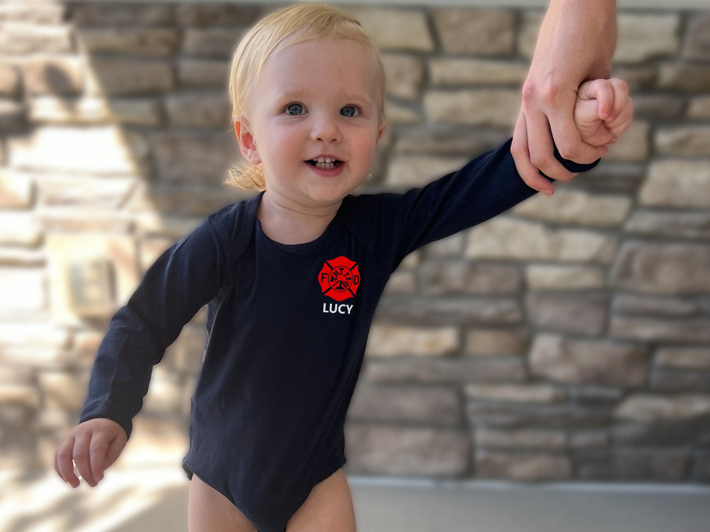 Blonde baby wearing a long sleeve bodysuit with red Maltese cross on left chest and baby’s name underneath in white.