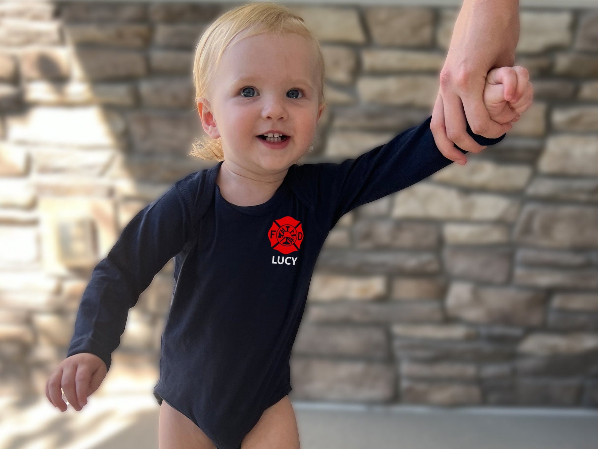 Blonde baby wearing a long sleeve bodysuit with red Maltese cross on left chest and baby’s name underneath in white.