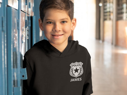 Black hooded sweatshirt for kids features a custom police badge design with a star and the text 'POLICE' above a name.  The name 'JAMES' is printed below the badge. The sweatshirt is shown against a blue locker background.