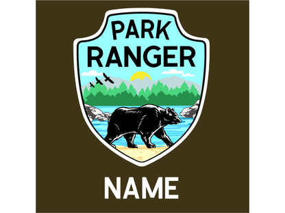 A blue and white shield with the text 'PARK RANGER' in black. The shield has a black bear walking on a beach with green trees and a yellow sun in the background. The text 'NAME' is below the shield.