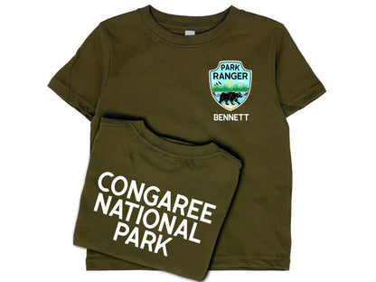 Dark green t-shirt front and back. Front shows a park ranger badge on left chest with mountains, water and black bear and a name written underneath in white. Back shows custom national park name in white.