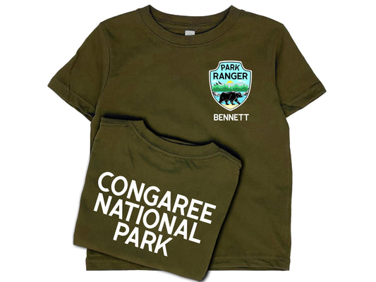Dark green t-shirt front and back. Front shows a park ranger badge on left chest with mountains, water and black bear and a name written underneath in white. Back shows custom national park name in white.