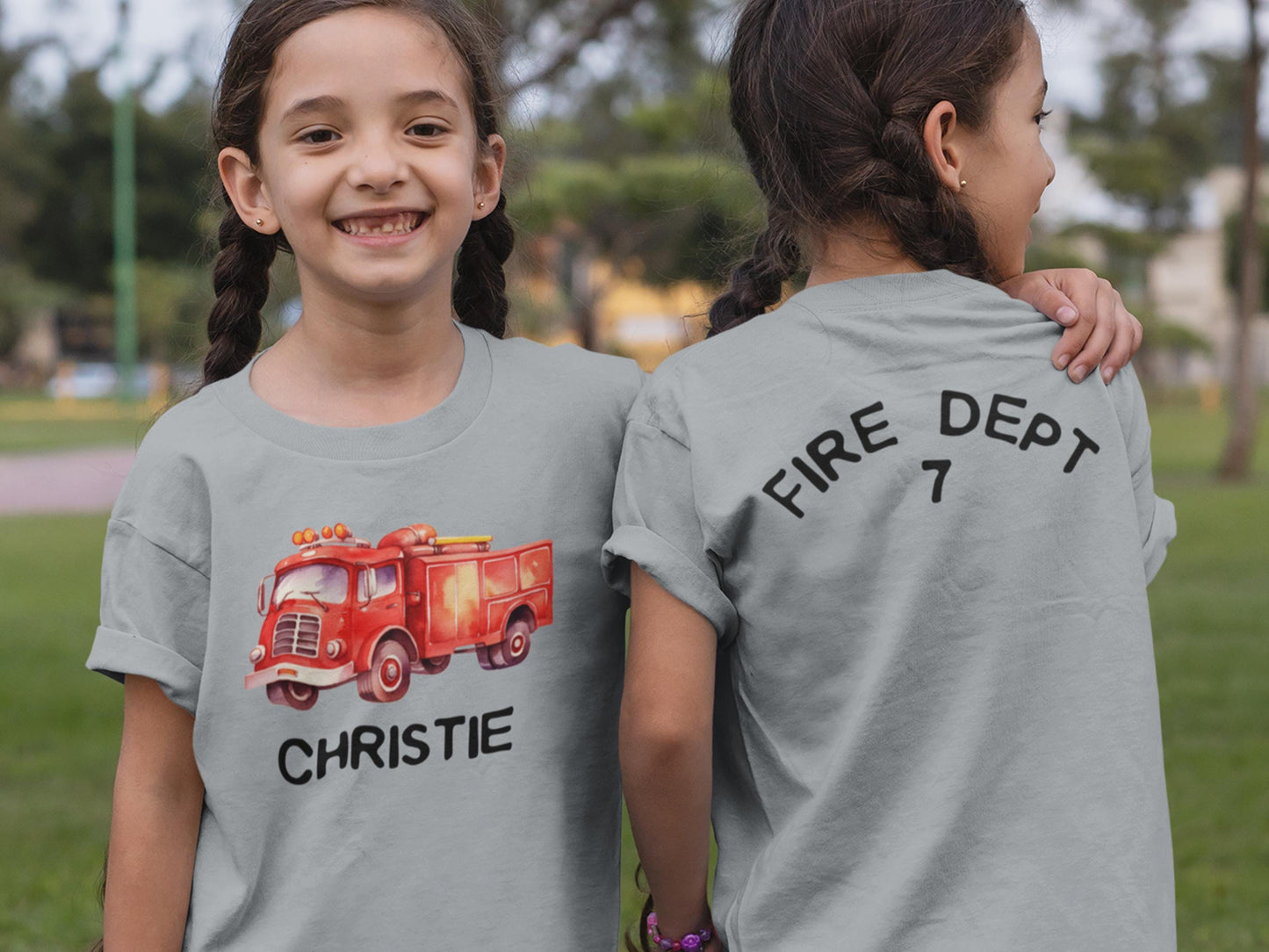 Twin girls wearing gray t-shirts, one facing forward with a red vintage watercolor firetruck printed on chest and the name Christie underneath. The other twin is facing backward with a large number 7 printed on the shirt.
