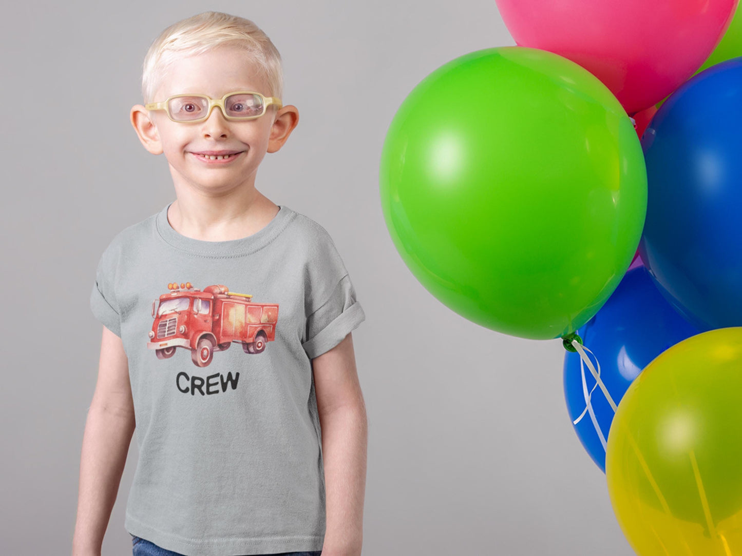 Young boy with glasses and smiling wearing a gray short sleeve t-shirt with a red vintage watercolor firetruck on the front chest. The name Crew is printed under the fire truck. Colorful balloons are to his left.