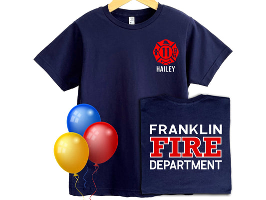 Navy blue kids’ t-shirt with a red firefighter Maltese cross on the front left chest, customizable with an age and name (shown as “11” and “Hailey”). The back of the shirt displays bold white and red text that can be personalized with any fire department name (shown as “FRANKLIN FIRE DEPARTMENT”). Three colorful balloons—yellow, red, and blue—add a festive touch. Perfect for birthdays or gifts for young firefighter fans.