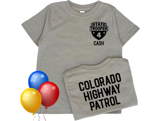 A gray toddler t-shirt with a black state trooper patch on the front that says 'State Trooper' with a star on either side of the number '4' and the name 'Cash' below. The back of the shirt says 'Colorado Highway Patrol' in black text. Colorful balloons indicate a birthday theme.