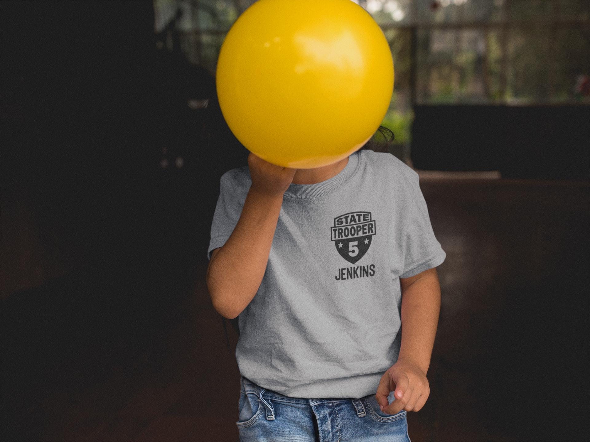 Toddler covering face with a balloon wearing a gray t-shirt with a black badge that says 'STATE TROOPER' and the number '5' with stars on each side. The name 'JENKINS' is underneath in black text.
