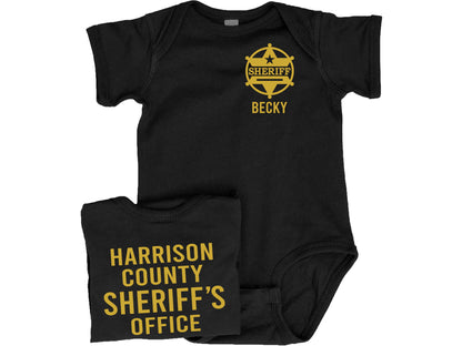 Black short sleeve baby bodysuit with gold sheriff badge on left chest and personalized name underneath. Back of bodysuit showing custom county sheriff’s office in gold text.