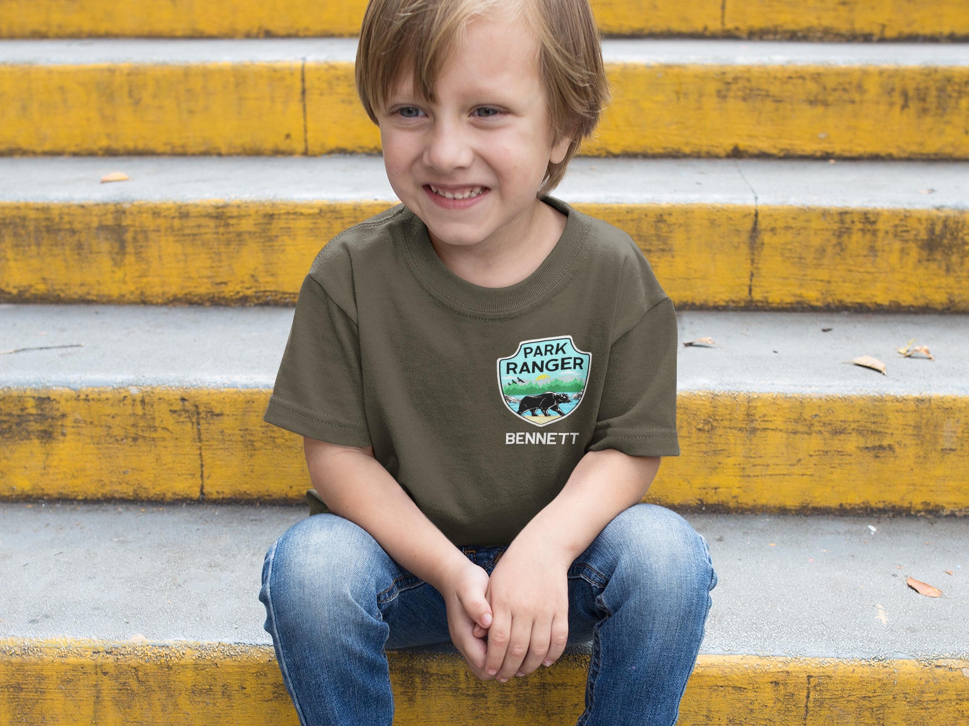 A young person wearing a green t-shirt with a patch that says 'Park Ranger' and 'Bennett' underneath. The patch has a green background with a brown bear and a blue sky with birds.