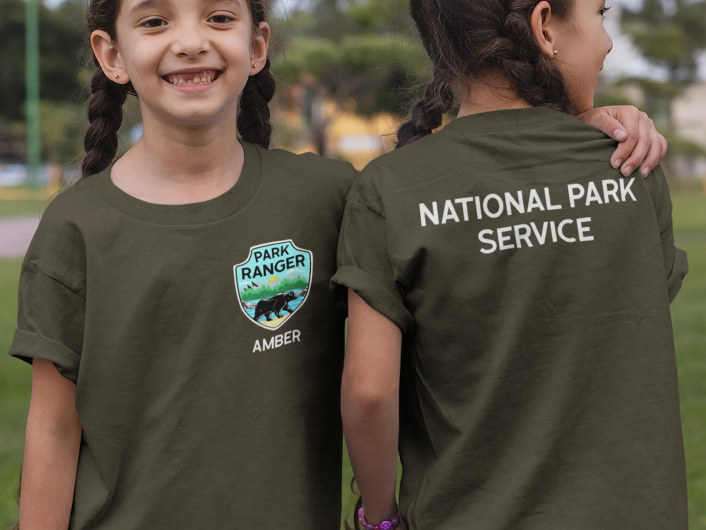 Two children wearing dark green t-shirts. The t-shirt on the left has a green, blue, and yellow patch with a bear and the text 'Park Ranger' and 'Amber'. The t-shirt on the right has the text 'National Park Service' on the back.