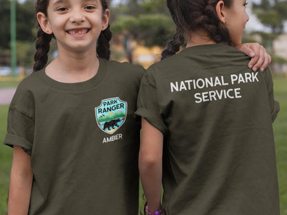 Two children wearing dark green t-shirts. The t-shirt on the left has a green, blue, and yellow patch with a bear and the text 'Park Ranger' and 'Amber'. The t-shirt on the right has the text 'National Park Service' on the back.
