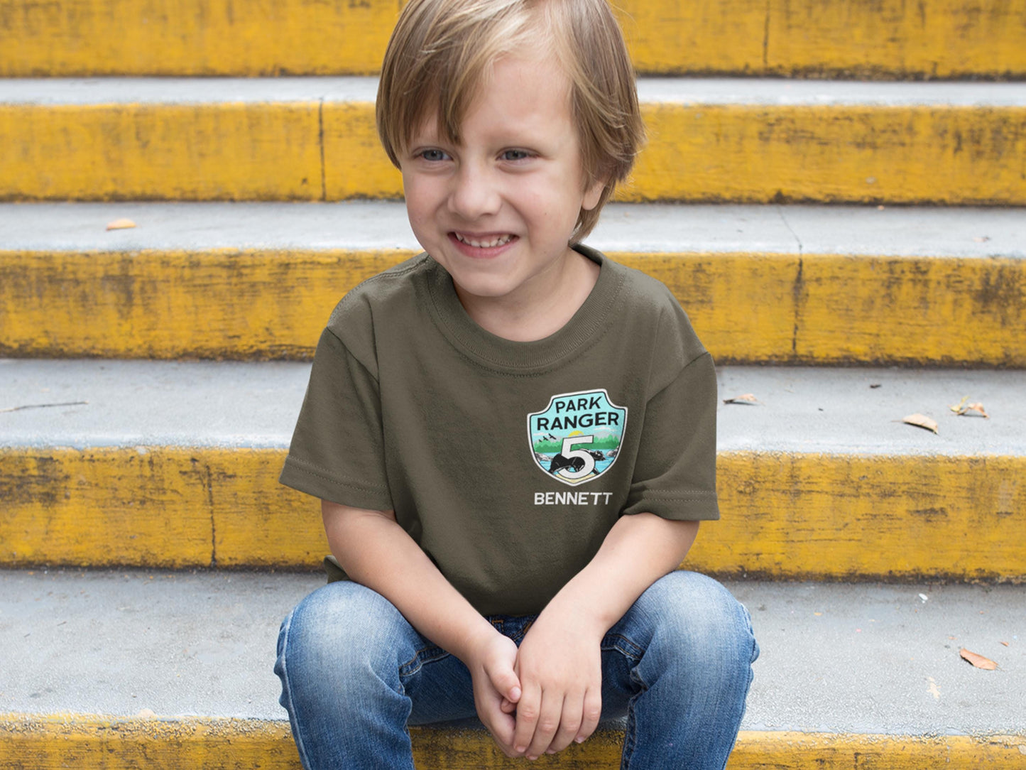 Smiling boy sitting on steps wearing a military green t-shirt. Shirt features a park ranger badge with the number 5 for birthday age and a name below the badge.