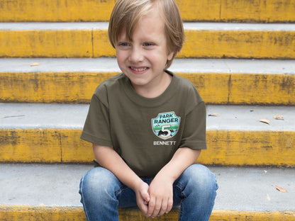 Smiling boy sitting on steps wearing a military green t-shirt. Shirt features a park ranger badge with the number 5 for birthday age and a name below the badge.