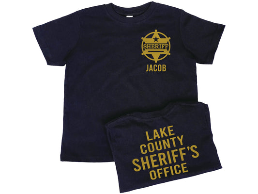 Black toddler t-shirt sporting a gold sheriff badge with 6 pointed star and a ring around it. The badge also has a 5 pointed star above the word Sheriff and the name "Jacob" underneath. The back of the shirt has gold lettering "Lake County Sheriff's Office" as a place holder for personalized Text.